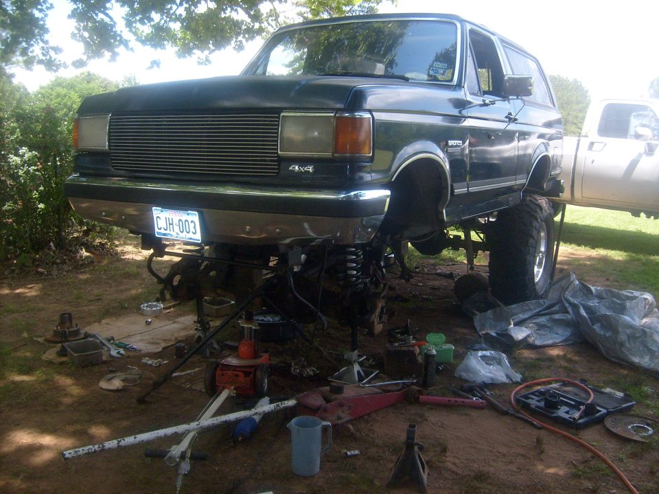 1989 Ford Bronco with front end disassembled, elevated on jack stands outdoors.