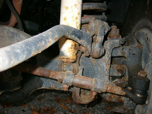 Rusty front suspension underside: white shock, sway bar, tie rod end and steering linkage on a 1987 Ford Bronco