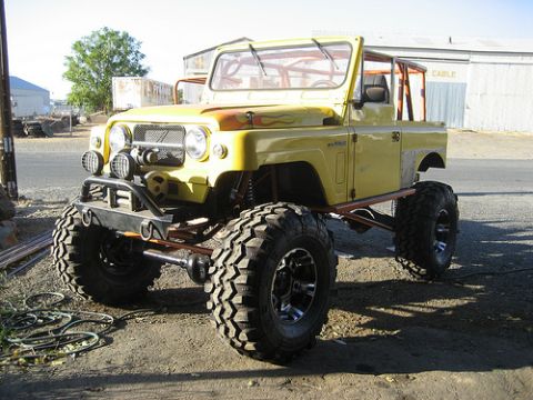1991 Ford Bronco - A heavily modified yellow off-road vehicle with oversized tires, a roll cage, and a winch is parked on a dirt lot in front of a building.