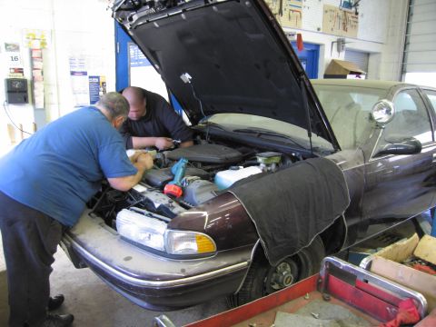 1991 Ford LTD Crown Victoria - Mechanics attend to the engine bay of a sedan, possibly a police car, with its hood raised.