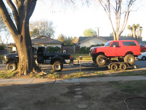 1987 Ford Bronco - A lifted red Ford Bronco is transported on a trailer by a black Ford pickup truck on a residential street.