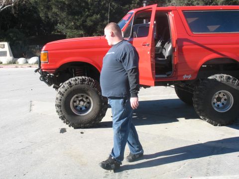 1987 Ford Bronco - A red 1987 Ford Bronco with significant suspension lift and large off-road tires is parked on a paved surface.