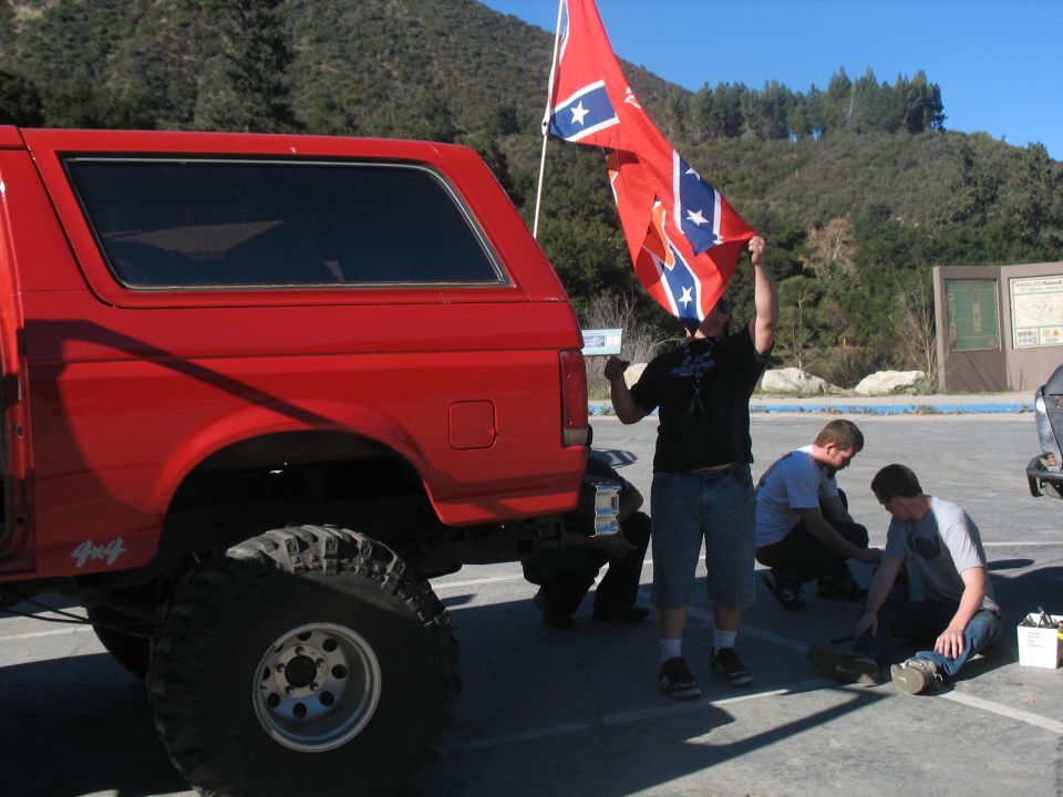 Red 1987 Ford Bronco parked, people gathered around, one holding a Confederate flag