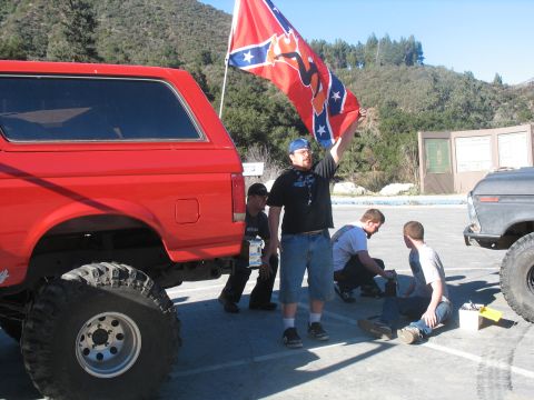 1987 Ford Bronco - A red Ford Bronco is parked in a lot with a group of people. One person is holding a Confederate flag, and another is working on something…