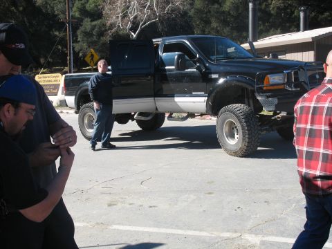 1987 Ford Bronco - A group of men stand around a lifted black Ford pickup truck with its driver's side door open. The truck has large off-road tires and a cus…