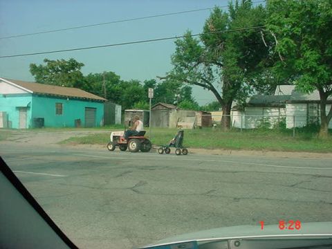1989 Ford Bronco - A person drives a lawn tractor with a trailer down a road past buildings and trees.