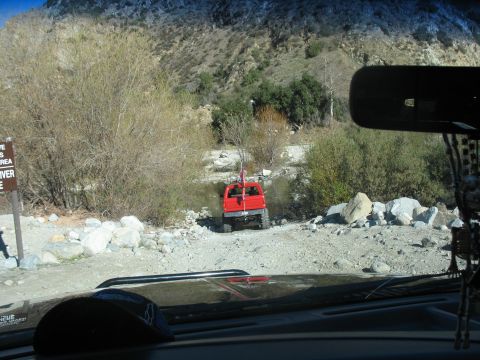 1987 Ford Bronco - A red off-road vehicle is parked near a riverbed with a mountainous, vegetated backdrop. The photo is taken from inside another vehicle, lo…