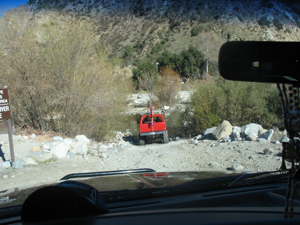Red off-road vehicle parked by a river in a mountainous area, viewed from inside another vehicle.