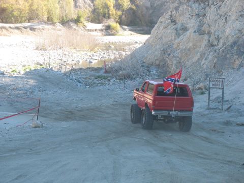 1987 Ford Bronco - A lifted red Ford Bronco with a Confederate flag drives on a dirt road next to a rocky hillside. A sign indicating "CROSS HERE" is visible…