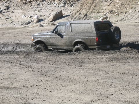 1987 Ford Bronco - A mud-covered, dark-colored SUV is stuck in thick mud, with its tires submerged. The vehicle has a spare tire mounted on the rear.