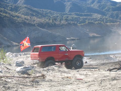 1987 Ford Bronco - A lifted red Ford Bronco with a Confederate flag is parked on a dirt and rocky shore next to a body of water with mountains in the backgrou…