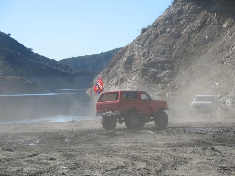 1987 Ford Bronco - A lifted, red Ford Bronco with a Confederate flag is driving through mud and dust near a body of water with a white truck in the background.