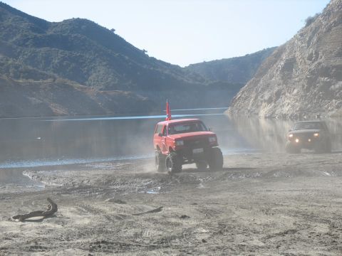 1987 Ford Bronco - A red Ford Bronco is driving through mud near a lake, kicking up dust. Another dark-colored SUV is visible in the background.
