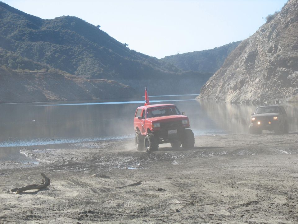Red Ford Bronco driving through mud near a lake, kicking up dust.