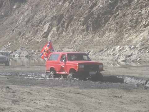 1987 Ford Bronco - A red Ford Bronco is driving through mud with a Confederate flag attached to its antenna. Another white SUV is visible in the background.