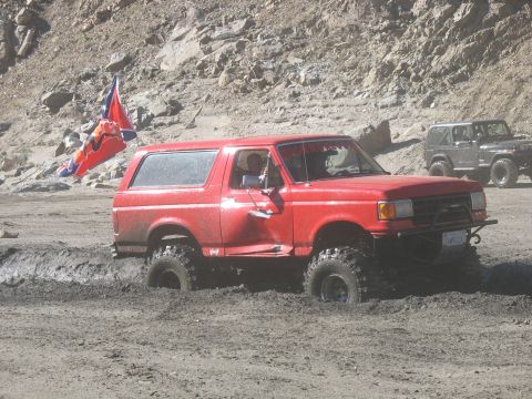 1987 Ford Bronco - A red Ford Bronco is stuck in thick mud, with a flag attached to the rear. Another Jeep is visible in the background.