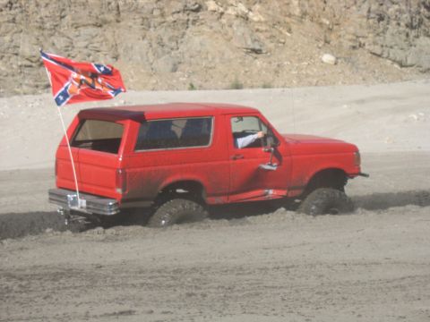 1987 Ford Bronco - A red Ford Bronco is stuck in the mud with a Confederate flag flying from a pole on the rear. The vehicle appears to be an off-road vehicle…