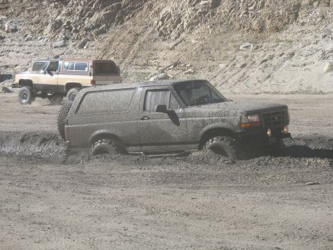 1987 Ford Bronco - A dark-colored Ford Bronco is stuck in thick mud, with its tires submerged. Another tan and brown SUV is visible in the background on a dir…