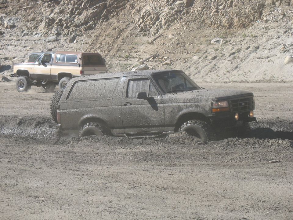 Mud-covered Ford Bronco stuck in mud, with another SUV in the background.