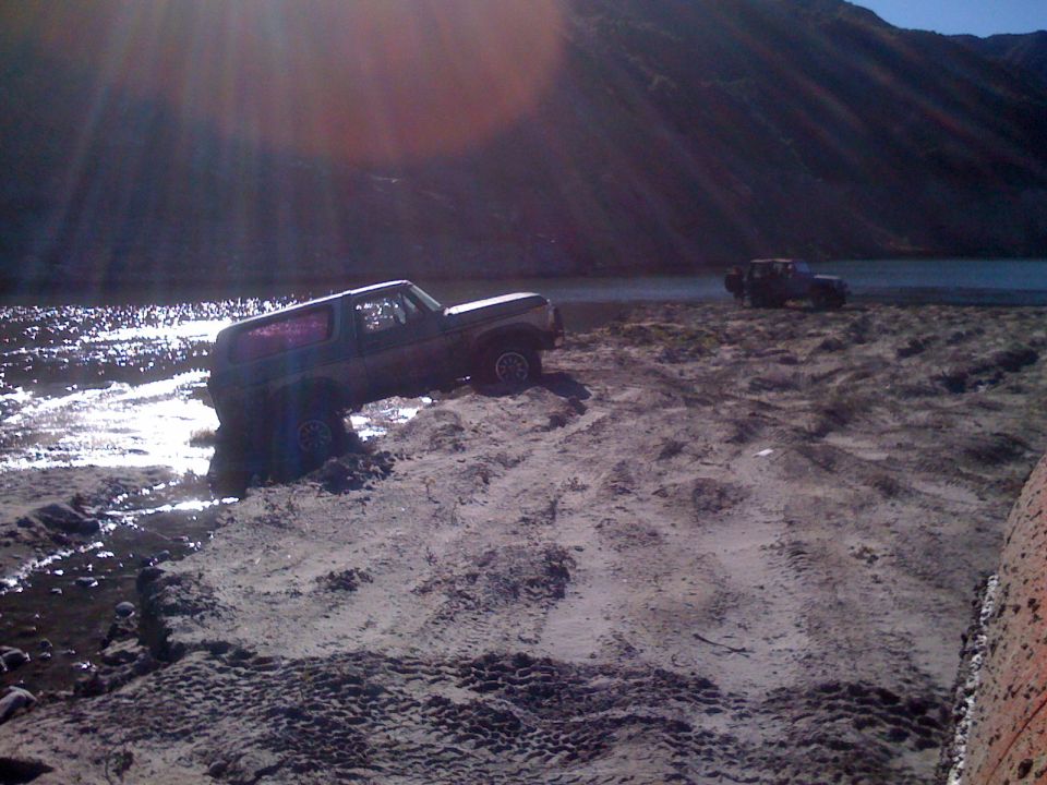 Dark Ford Bronco stuck in mud and water on a sandy bank.