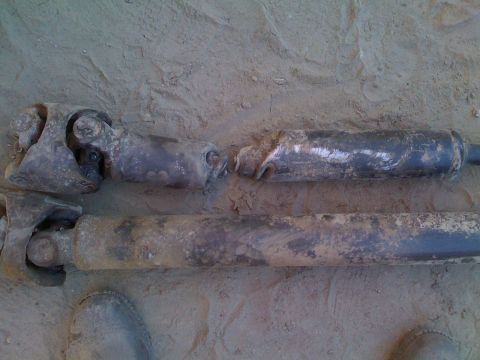 1979 Ford Bronco - Two driveshafts, one of which appears to be damaged, lie on the sandy ground. The driveshafts are covered in dirt and mud.