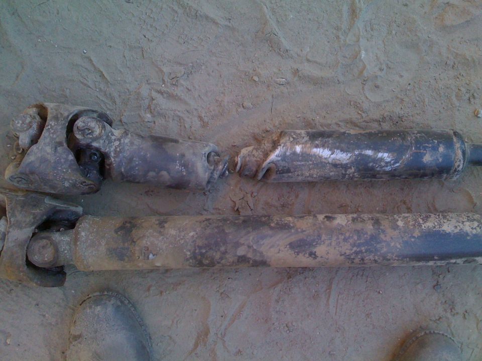 Two dirty driveshafts on sandy ground, one appears damaged.