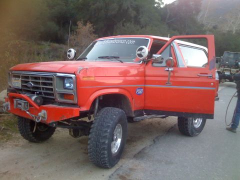 1979 Ford Bronco - A bright orange, lifted Ford Bronco with its driver's side door open is parked on a dirt road with trees and hills in the background. The v…