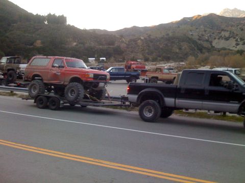 1979 Ford Bronco - A lifted red Ford Bronco is being towed on a trailer by a black Ford F-series pickup truck. Several other trucks and SUVs are parked in the…
