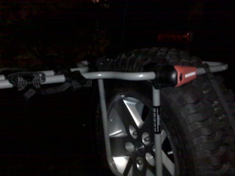 2008 Jeep Wrangler - Night shot of a silver spare-tire-mounted bike rack strapped to the rear-mounted spare of a vehicle, with bungee cords and the rack's black…