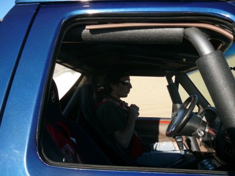 1988 Ford Bronco - Side view of the interior of a blue Ford Bronco, showing a person wearing sunglasses and a racing harness. The vehicle appears to be in a s…