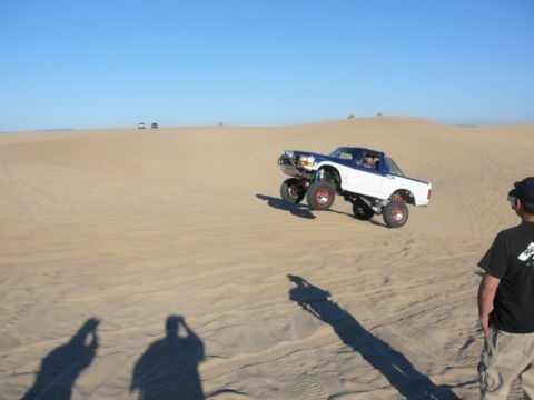 1988 Ford Bronco - A modified white and blue pickup truck with large tires is shown airborne in a desert landscape. A person wearing a black shirt and hat is…