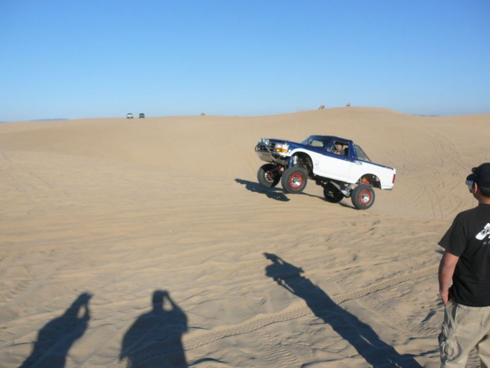 White and blue pickup truck jumping in the desert with a person watching.