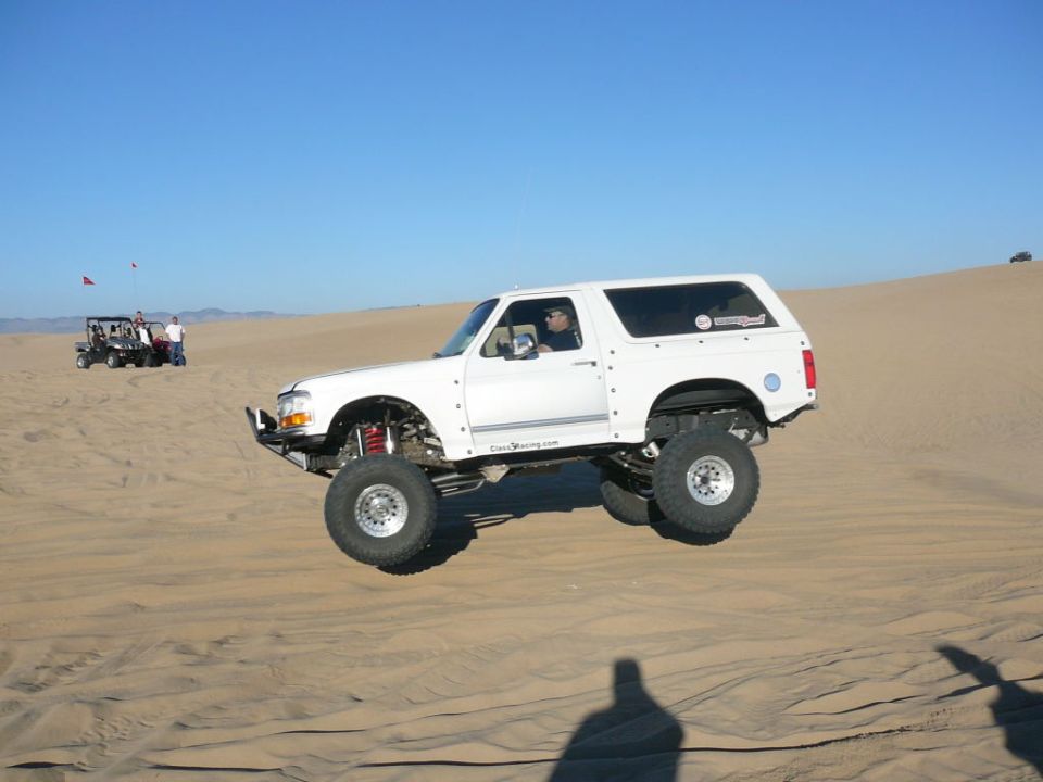 White lifted Ford Bronco jumping over a sand dune with a clear blue sky.