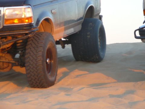 1988 Ford Bronco - The front section of a lifted Ford Bronco is shown in profile, with its large off-road tires resting on sand dunes. The vehicle's headlight…