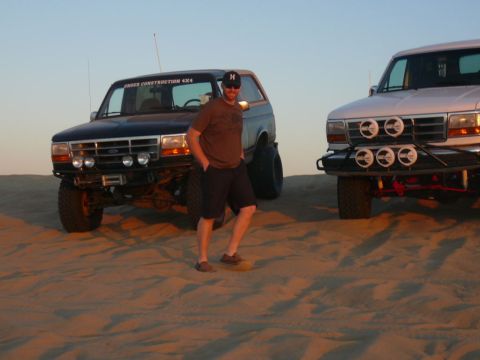 1988 Ford Bronco - A man stands between two modified Ford Broncos in a sandy, desert-like environment during sunset. The Bronco on the left is black and has "…