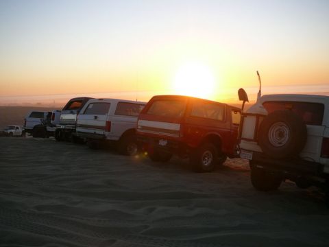 1988 Ford Bronco - Several Ford Broncos are parked in a line on a sand dune at sunset. The vehicles are facing towards the right of the frame, with the sun se…