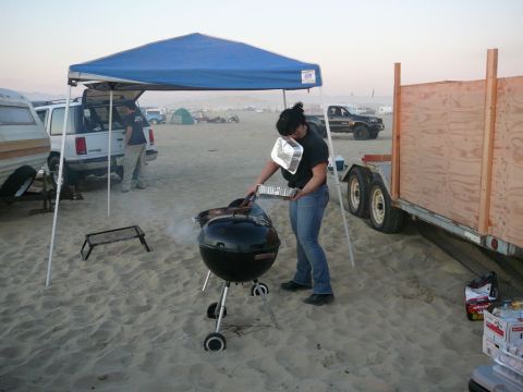 1988 Ford Bronco - A woman is grilling food on a charcoal grill on a sandy beach, with a blue canopy set up over a white SUV and a trailer in the background.