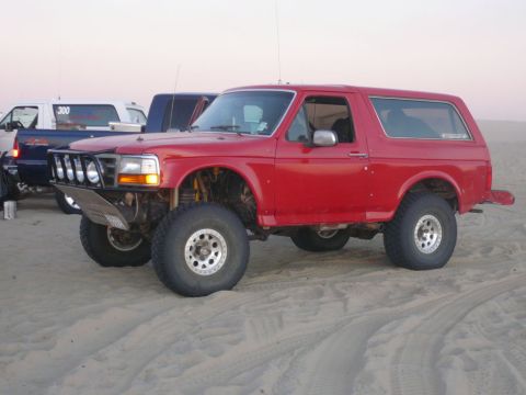 1988 Ford Bronco - A red 1988 Ford Bronco with off-road modifications is parked on a sandy beach. The vehicle features a custom front bumper with four lights…