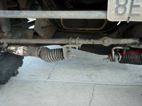1995 Ford Bronco - A close-up view of the steering linkage of a vehicle, showing a damaged tie rod end with a torn rubber boot and a red steering stabilizer s…