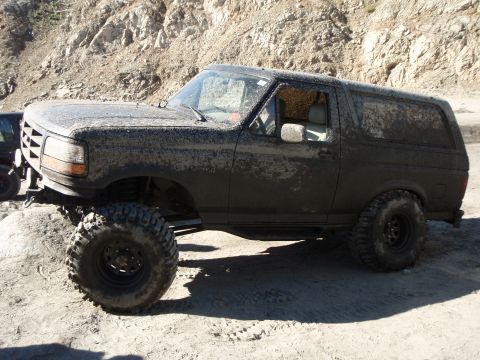 1995 Ford Bronco - A dark-colored, mud-covered Ford Bronco is parked on a sandy, rocky terrain with a steep hillside in the background. The vehicle has large…