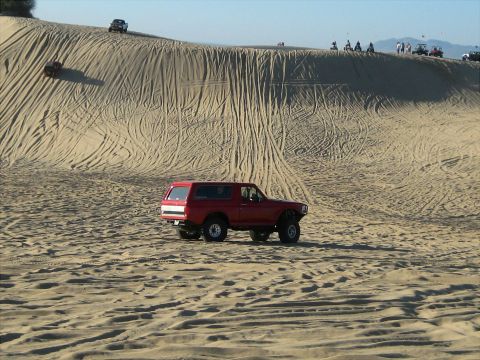 1996 Ford Bronco - A red Ford Bronco is parked on a sandy dune with tire tracks visible on the slope behind it. Several other off-road vehicles and people are…