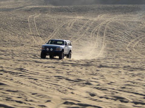 1996 Ford Bronco - A white Ford Bronco is driving on a sand dune, kicking up dust. The vehicle has aftermarket lights and a modified bumper.