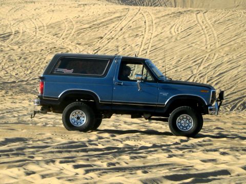1996 Ford Bronco - A blue Ford Bronco is driving on sand dunes, kicking up dust. The vehicle is seen from the side, with its front left wheel prominently disp…