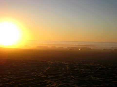 1996 Ford Bronco - The sun is setting over a beach with sand dunes in the foreground and buildings in the distance.