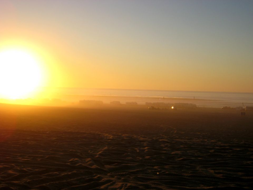 Sunset over a beach with sand dunes and distant buildings.