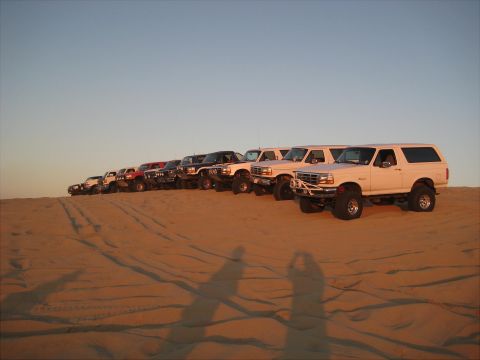 1996 Ford Bronco - A line of modified Ford Broncos are parked on a sand dune under a clear sky. The vehicles are angled towards the right, with the closest on…