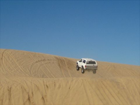 1996 Ford Bronco - A white 1996 Ford Bronco is captured mid-air as it jumps over a sand dune under a clear blue sky. The vehicle is seen from the rear-three-q…