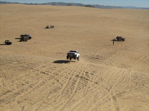 1996 Ford Bronco - A white Ford Bronco is captured mid-air as it jumps over a sand dune. Multiple other off-road vehicles are visible in the sandy desert land…
