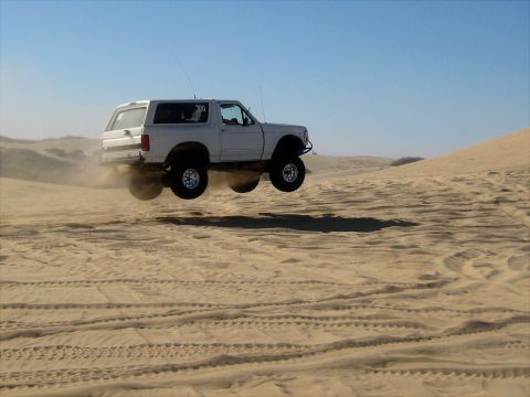 1996 Ford Bronco - A white Ford Bronco is captured mid-air as it jumps over sand dunes, kicking up dust. The SUV is viewed from the side against a clear blue…