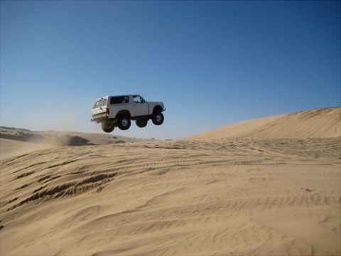 1996 Ford Bronco - A white Ford Bronco is captured mid-air, jumping over a sand dune under a clear blue sky.